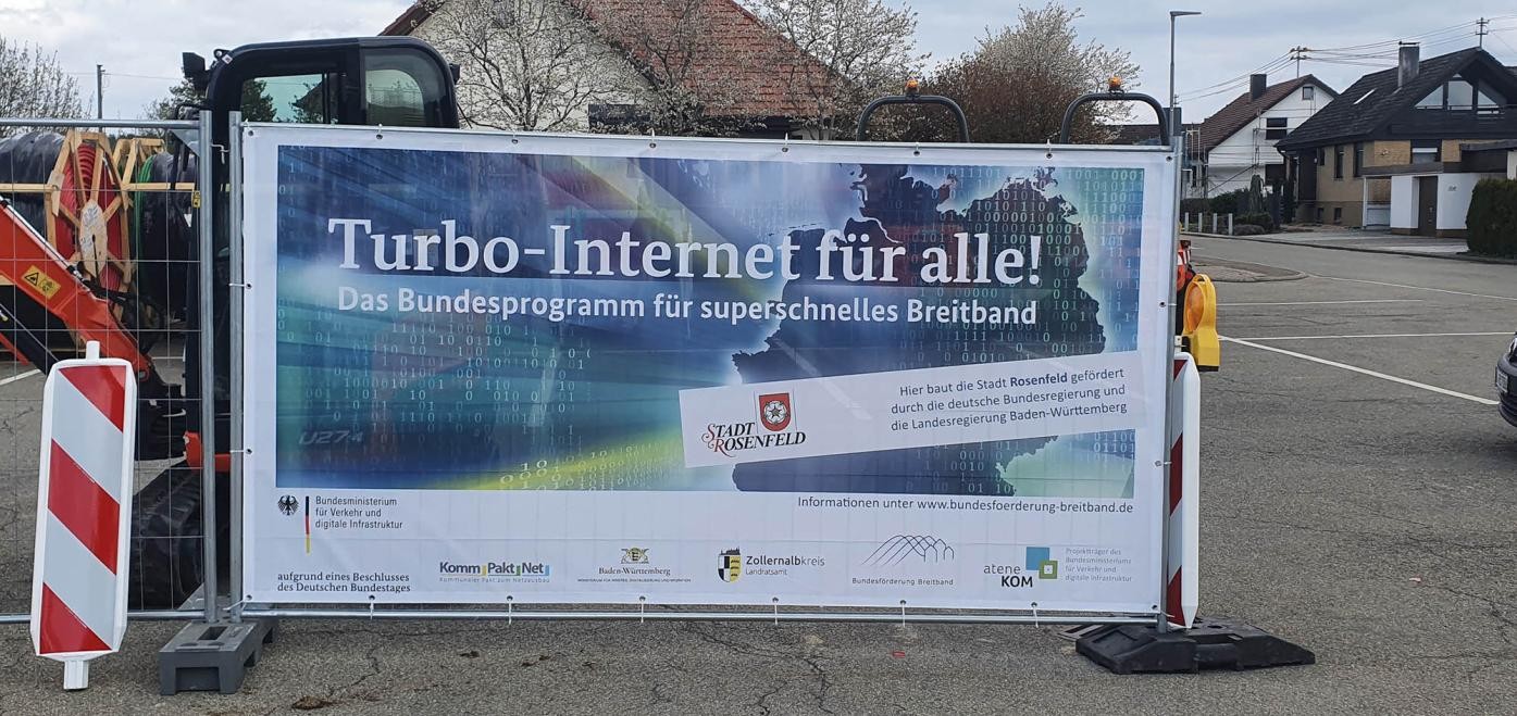 Zusehen ist ein Bauzaun mit einem Plakat mit der Aufschrift "Turbo-Internet für alle!" rechts und links davon steht jeweils eine Pylone